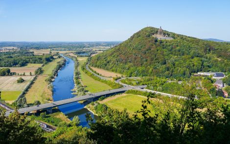 Weite Flusslandschaft mit Feldern und Brücken, im Vordergrund ein bewaldeter Hügel mit einem Denkmal auf der Spitze, aufgenommen aus erhöhter Perspektive.