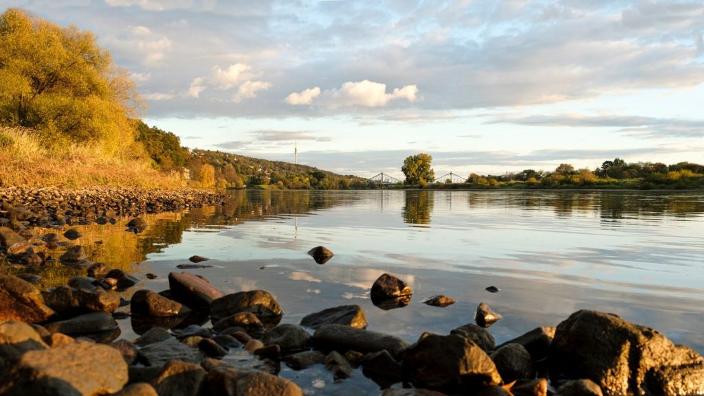 Ruhige Flusslandschaft mit Ufersteinen im Vordergrund, reflektierendem Wasser und einer Brücke in der Ferne unter bewölktem Himmel. Goldene Herbstfarben am Ufer.