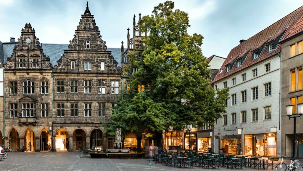 Historischer Platz in Münster, Deutschland, mit verzierten Gebäuden, Brunnen und einem großen Baum. Cafés laden zum Verweilen ein. Münsterland entdecken!