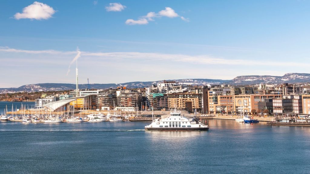 Oslo Fjord mit Fähre vor der Skyline. Moderne Architektur, Yachthafen und Berge im Hintergrund unter blauem Himmel. Norwegens Hauptstadt.