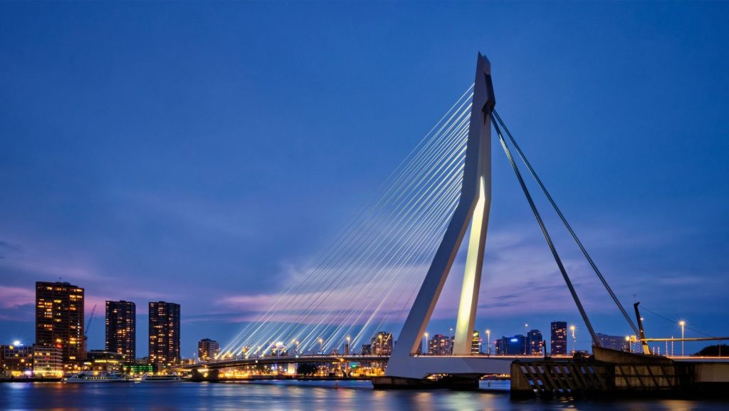Die Erasmusbrücke in Rotterdam erstrahlt im Abendlicht. Das Wahrzeichen der Stadt mit seinen markanten Pylonen und Stahlseilen spiegelt sich im Wasser. Im Hintergrund die Skyline von Rotterdam bei Nacht.