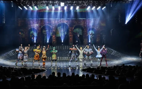 Szene aus "Starlight Express" in Bochum: Darsteller in farbenfrohen Rollschuhkostümen auf der Bühne, beleuchtet von Scheinwerfern, vor einer Backsteinmauer-Kulisse.