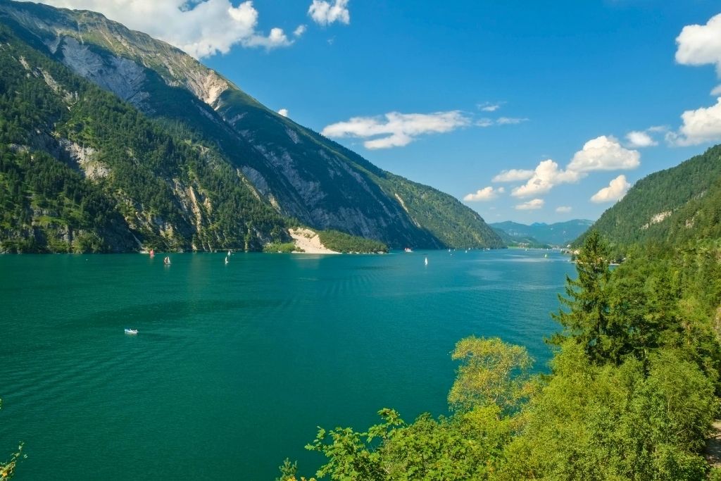 Türkisblauer Achensee in Tirol, umrahmt von bewaldeten Bergen unter blauem Himmel mit Wolken. Segelboote gleiten über das Wasser. Im Hintergrund die Zillertaler Alpen.