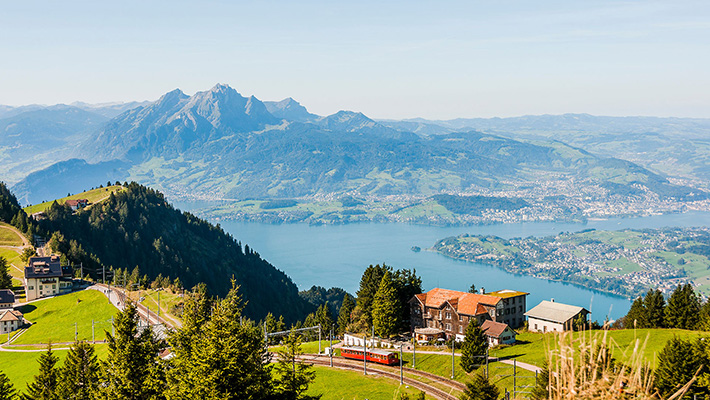 Panoramablick auf den Vierwaldstättersee in der Schweiz mit dem Pilatus im Hintergrund. Eine rote Zahnradbahn schlängelt sich durch die grüne Landschaft.
