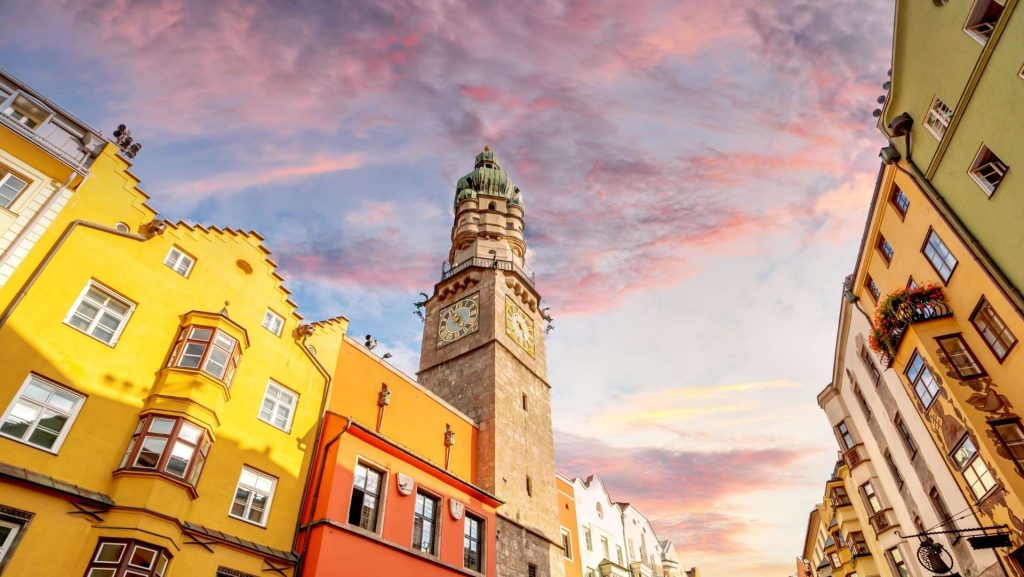 Innsbrucks Stadtturm ragt vor einem rosafarbenen Himmel empor, umgeben von farbenfrohen Häusern. Ein Panorama, das zum Durchatmen einlädt – besondere Momente in Europa.