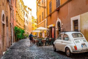 Gasse in Rom mit Kopfsteinpflaster, einem weißen Kleinwagen und einem Straßencafé mit Sonnenschirmen. Warme Farben und südländisches Flair.