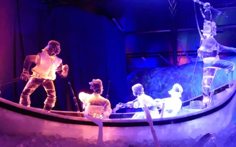 Eisskulpturen: Eine Szene mit Figuren in einem Eisboot, beleuchtet in Blau und Lila. Detailreiche Eisfiguren stellen Seeleute dar, die in einem eisigen Ozean fahren.