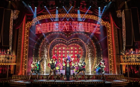 Bühnenbild des Musicals „Moulin Rouge“ in Köln: Rotes Bühnenbild mit Herz, Tänzern und dem leuchtenden Schriftzug „Moulin Rouge“.