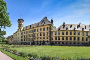 Schlossgarten des Stadtschlosses Fulda mit gelbem Schloss vor blauem Himmel. Blumen und Rasenflächen im Vordergrund.