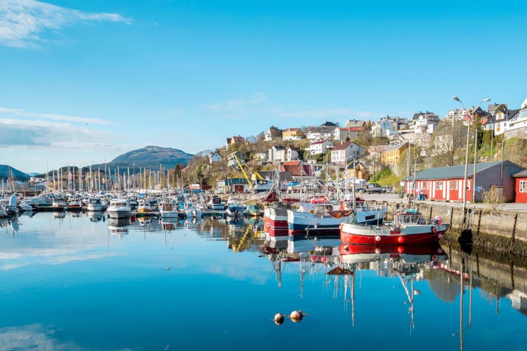 Malerischer Hafen in Kristiansund, Norwegen mit bunten Booten, die sich im ruhigen, blauen Wasser spiegeln. Im Hintergrund erstreckt sich eine Hügellandschaft mit Häusern unter einem strahlend blauen Himmel.