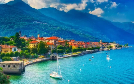 Malerische Küstenstadt in Italien mit bunten Häusern am Comer See, umgeben von grünen Bergen unter blauem Himmel. Segelboote ankern im türkisfarbenen Wasser.