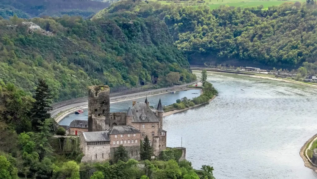 Majestätische Burg thront über dem Rhein, eingebettet in grüne Hänge. Der Rhein schlängelt sich durch die Hänge in die Ferne.