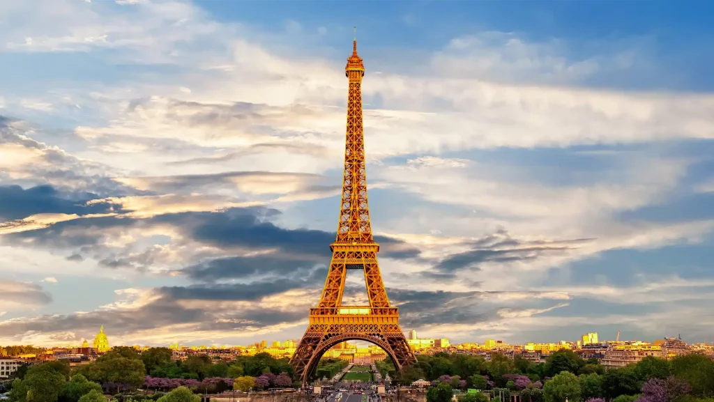 Die goldene Silhouette des Eiffelturms in Paris erstrahlt vor einem dramatischen Himmel mit Wolken. Im Vordergrund grüne Bäume und die Stadtlandschaft der Weltmetropole.