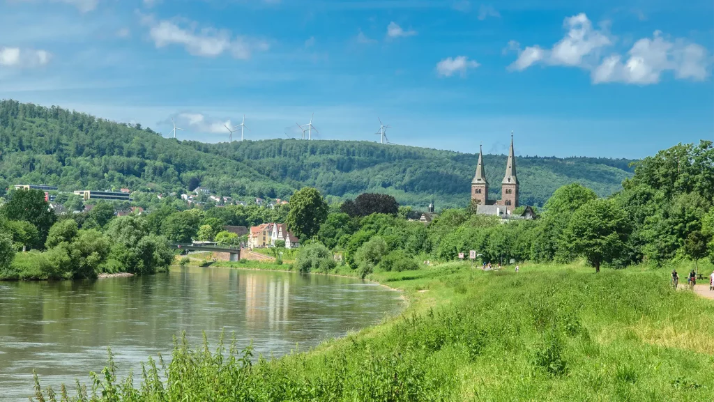 Weser-Flusslandschaft in Deutschland mit grünen Ufern, einer Stadt im Hintergrund und Windrädern auf einem bewaldeten Hügel. Zwei Kirchtürme ragen über die Stadt hinaus.