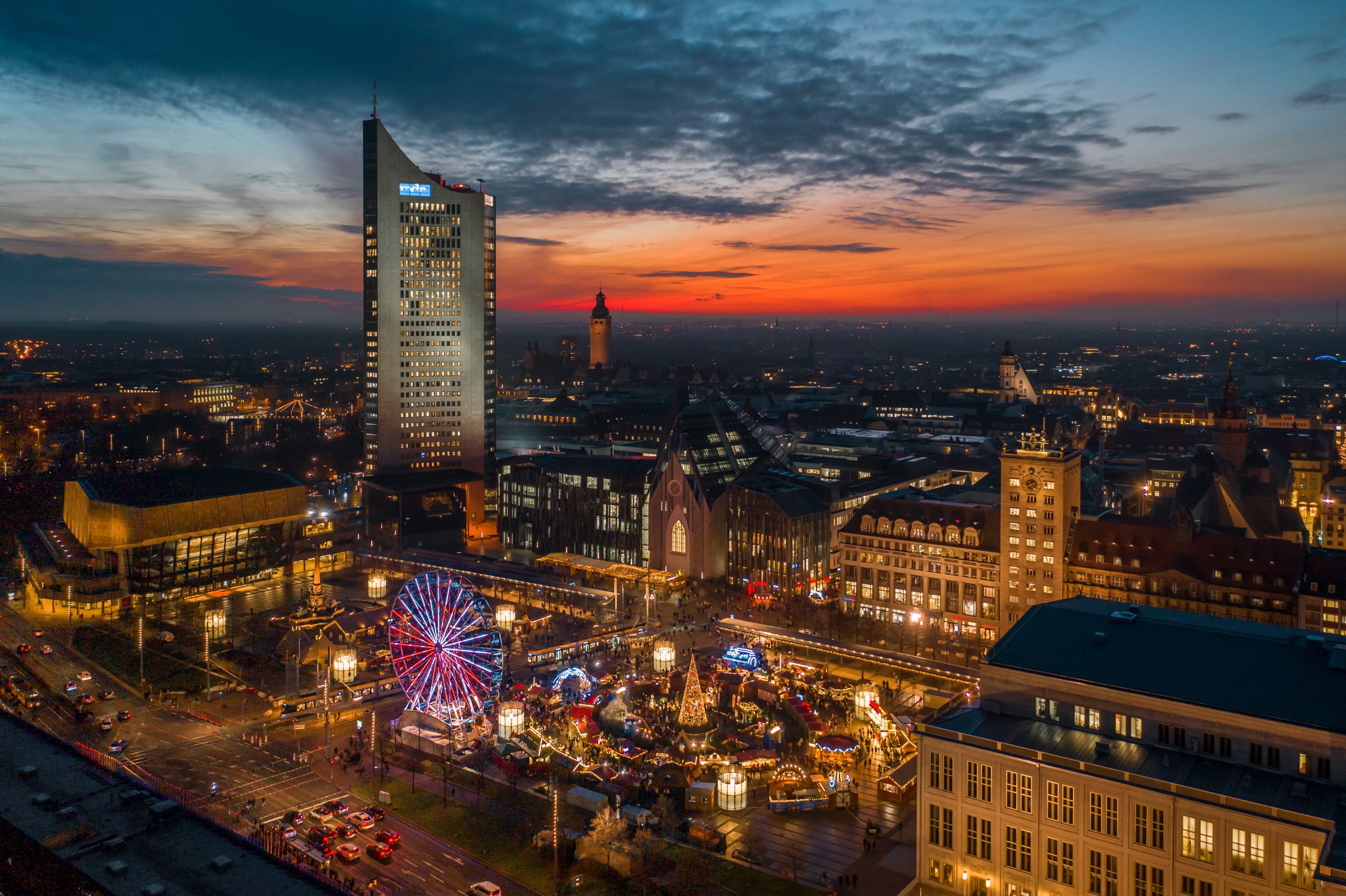 Luftaufnahme des Leipziger Weihnachtsmarktes bei Sonnenuntergang, mit beleuchtetem Riesenrad, Buden, dem Gewandhaus und dem City-Hochhaus im Hintergrund.
