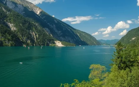 Türkisblauer Achensee, eingebettet zwischen bewaldeten Bergen unter blauem Himmel mit Wolken. Segelboote und ein kleines Boot auf dem See.