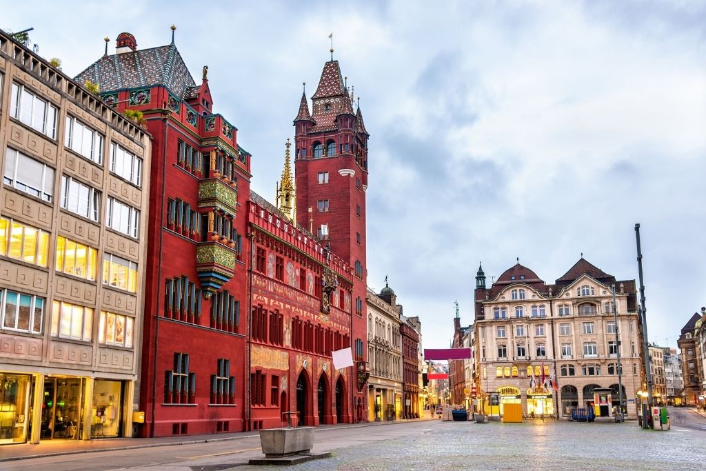 Das Basler Rathaus, ein markantes rotes Gebäude, dominiert den Marktplatz in Basel, Schweiz. Architekturdetails und ein bewölkter Himmel erzeugen eine malerische Szene in der Schweiz.