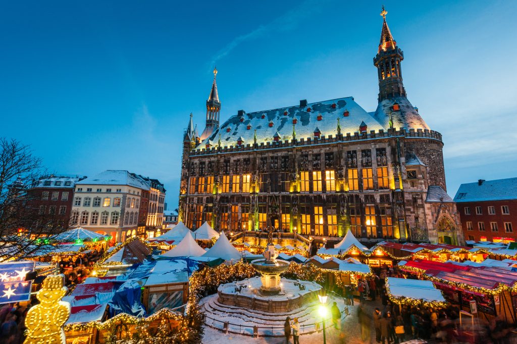 Festlicher Weihnachtsmarkt vor dem Aachener Rathaus im Dreiländereck, beleuchtet bei Dämmerung mit Schnee und bunten Buden.