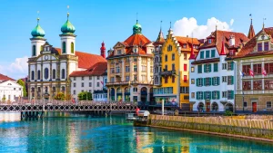 Bunte Häuserzeile in Luzern am Vierwaldstättersee mit markanter Jesuitenkirche. Spiegelung der Architektur im türkisfarbenen Wasser.