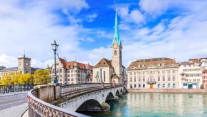 Zürichs Fraumünster Kirche mit grünem Turm, fotografiert vom gegenüberliegenden Ende einer Brücke über die Limmat. Blauer Himmel mit Wolken über der malerischen Altstadt.