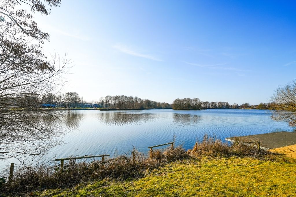 Ruhiger See im Münsterland unter blauem Himmel. Ufer mit Steg und grüner Wiese im Vordergrund, Bäume am Horizont. Ideal für Ausflüge.