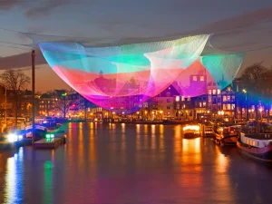 Bunt beleuchtete Netze hängen wie schwebend über dem Kanal in Amsterdam bei Abenddämmerung. Drumerhum liegen Boote an.
