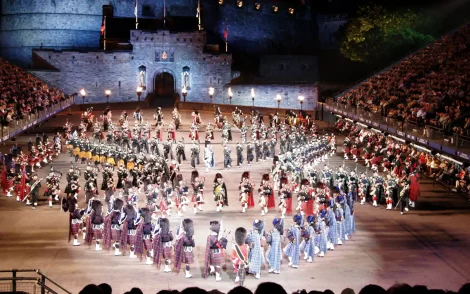 Nachtszene des Royal Edinburgh Military Tattoo vor dem erleuchteten Edinburgh Castle. Dudelsackspieler und Tänzer in traditionellen schottischen Trachten treten vor einer großen Zuschauermenge auf.