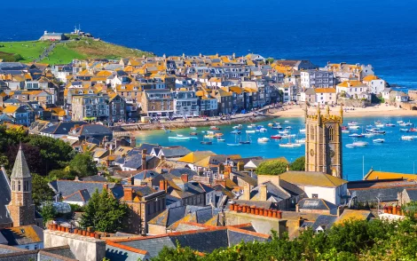 Malerische Aussicht auf St Ives, Cornwall, mit Häusern mit orangefarbenen Dächern, einem Hafen voller Boote und einer Kirche mit Turm.