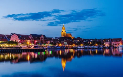Abendliche Silhouette von Waren an der Müritz mit beleuchteten Gebäuden, einem Kirchturm und Spiegelungen auf dem See.