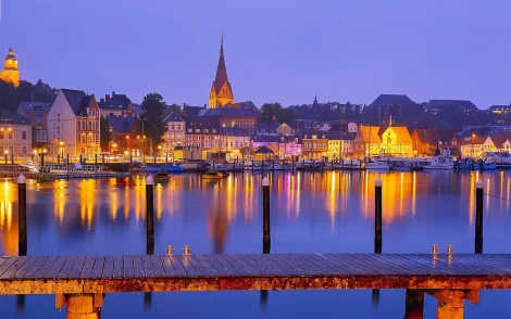 Abendliche Ansicht einer norddeutschvon Flensburg mit beleuchteten Gebäuden und Spiegelungen im Wasser, ein Steg im Vordergrund.