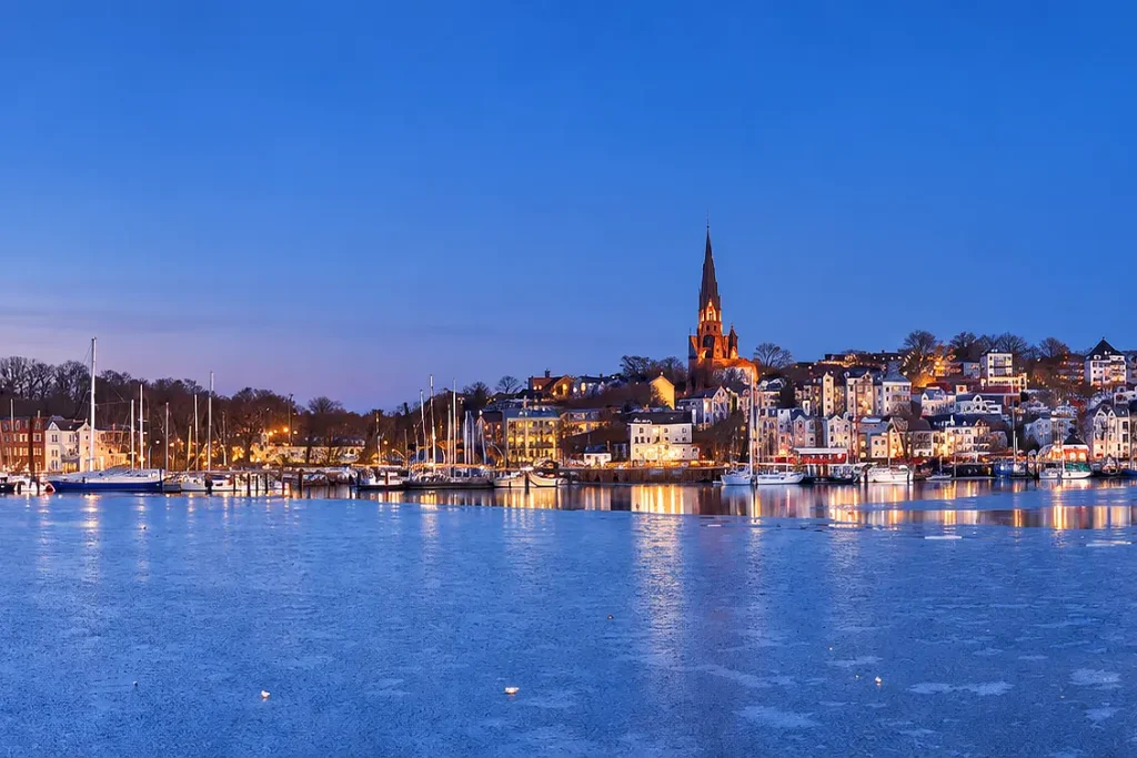 Flensburger Hafen bei Dämmerung mit beleuchteten Gebäuden, Segelbooten und einer Kirche im Hintergrund, Adventserlebnis im Norden.