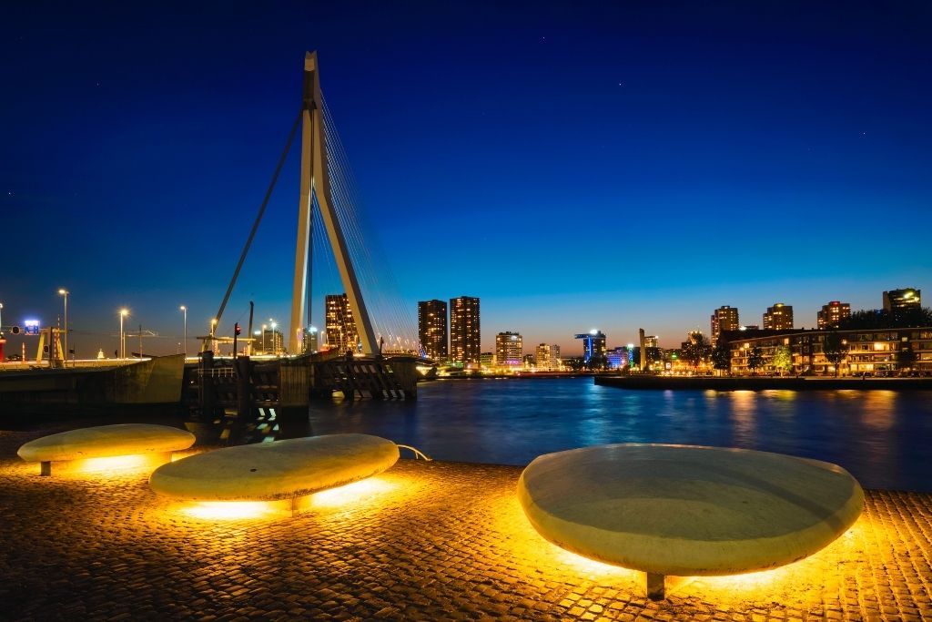 Rotterdam bei Nacht: Die Erasmusbrücke erstrahlt über der Nieuwe Maas, während beleuchtete Sitzgelegenheiten im Vordergrund zum Verweilen einladen. Perfekt für eine Wochenendreise zu den Rotterdamer Welthafentagen.