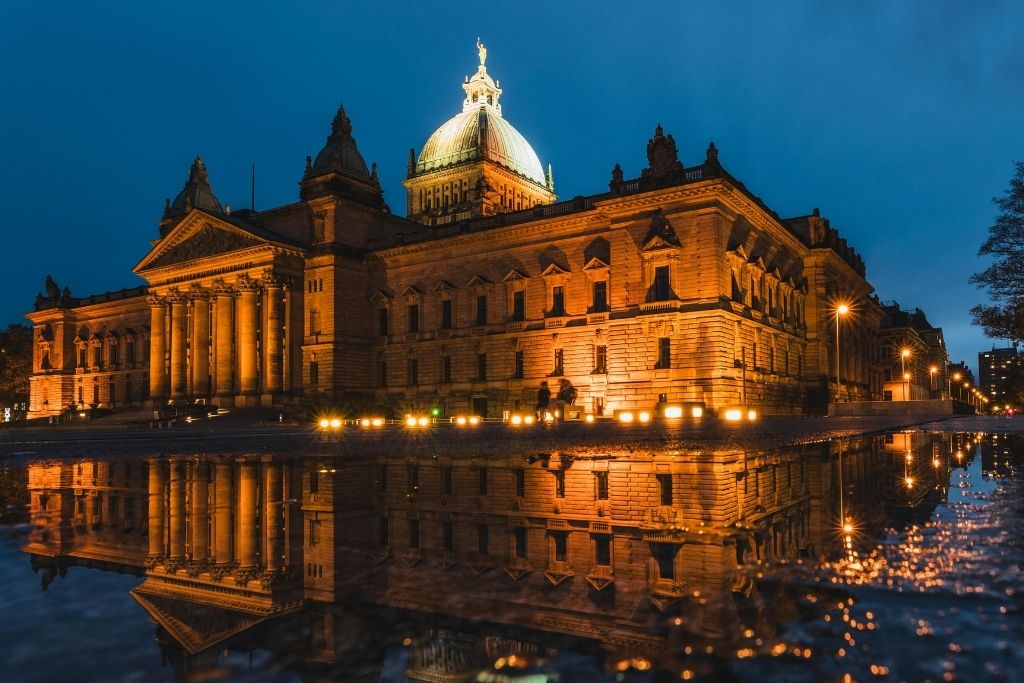 Leipzig bei Nacht: Das Neue Rathaus erstrahlt in warmem Licht, spiegelt sich in einer Pfütze auf dem Pflaster. Historische Architektur und moderne Beleuchtung vereint.