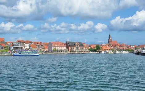 Blick über das Wasser auf Waren (Müritz) mit bunten Häusern, rotem Kirchturm und blauen Himmel. Boote liegen im Hafen.