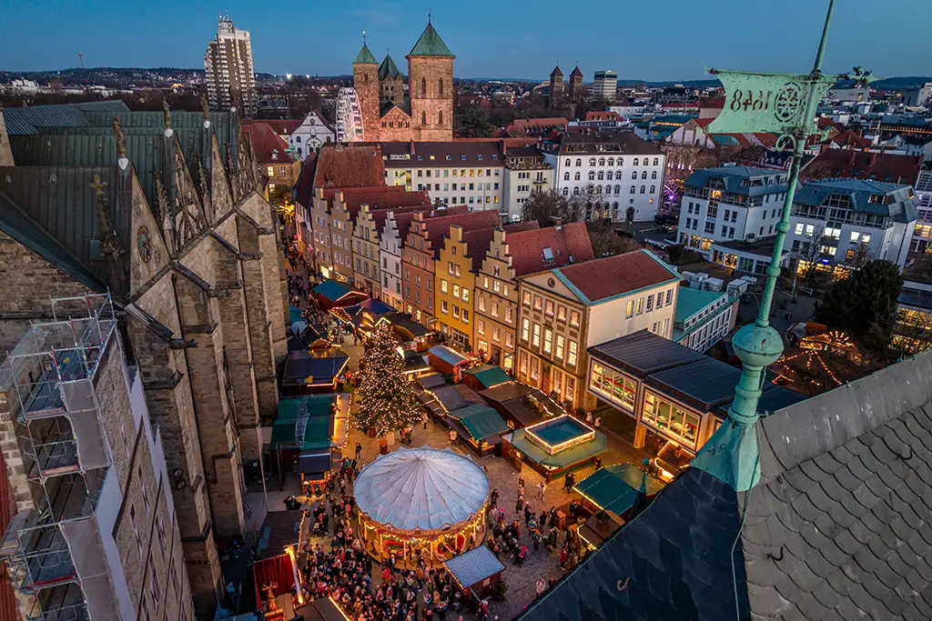 Ein festlich beleuchteter Weihnachtsmarkt in der Osnabrücker Innenstadt mit Karussell und Buden vor historischer Architektur bei Dämmerung.